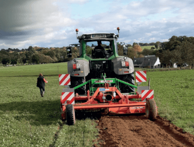 Paillage de haies et agroforesterie 1 Un tracteur vert laboure un champ, créant une bande de terre retournée pour le futur paillage. Une personne marche à proximité sur l'herbe. Des arbres et une maison blanche se dressent dans le paysage bocager sous un ciel partiellement nuageux.