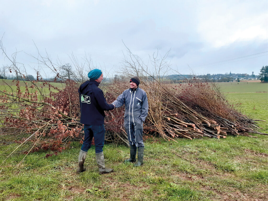Accompagnement à la gestion durable 2 Deux personnes, chaussées de bottes et de vêtements d'extérieur, se serrent la main dans un champ herbeux, avec un gros tas de bois et de branches en guise de paillage derrière elles. Le ciel nuageux et un village au loin complètent le paysage pittoresque du bocage en arrière-plan.