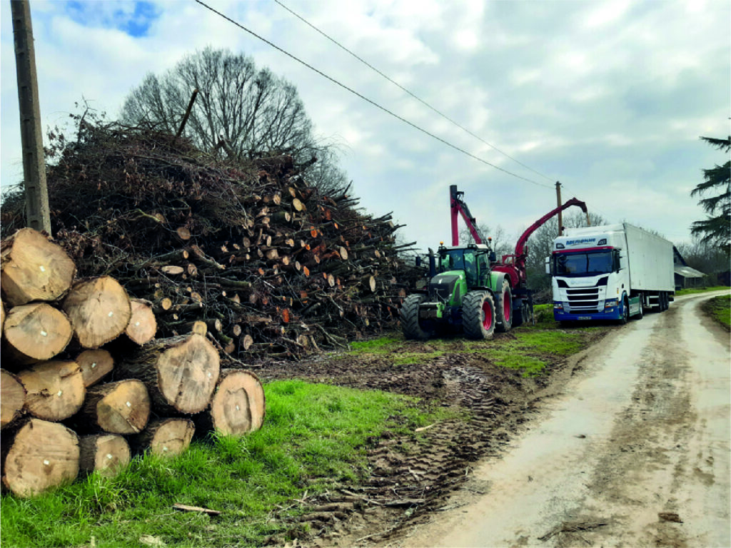 Un gros tas de bois coupé et de branches trône au bord d'une route rurale dans le bocage, avec un tracteur, un broyeur à bois et un camion blanc à proximité, sous un ciel nuageux. Des traces de pneus marquent le sol boueux, laissant entrevoir une production d'énergie continue.