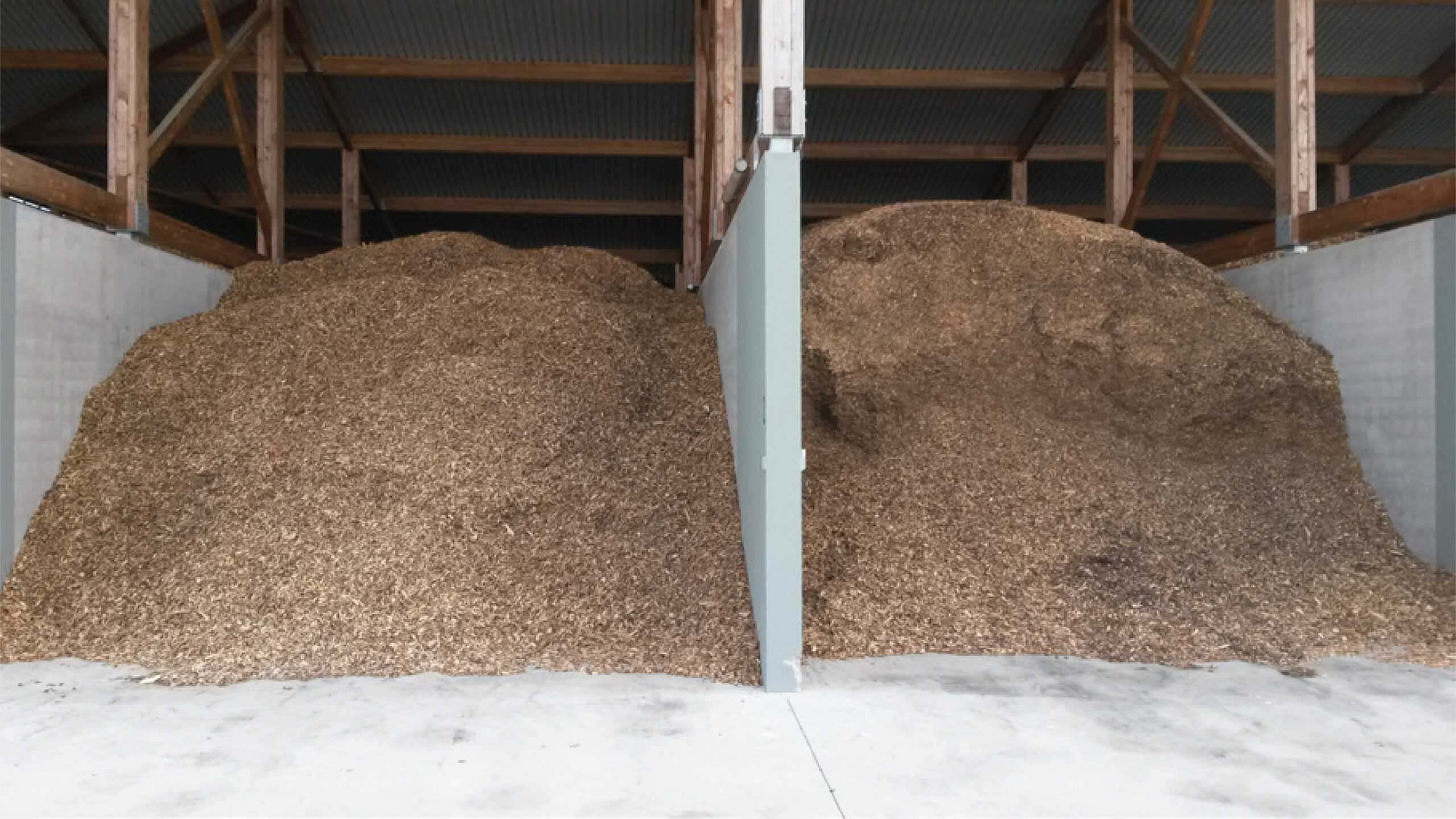 Vente de bois énergie 1 Deux grandes piles de copeaux de bois, idéales pour le paillage ou comme source d'énergie, sont stockées dans des sections séparées d'une installation intérieure avec des sols en béton et des poutres en bois robustes.